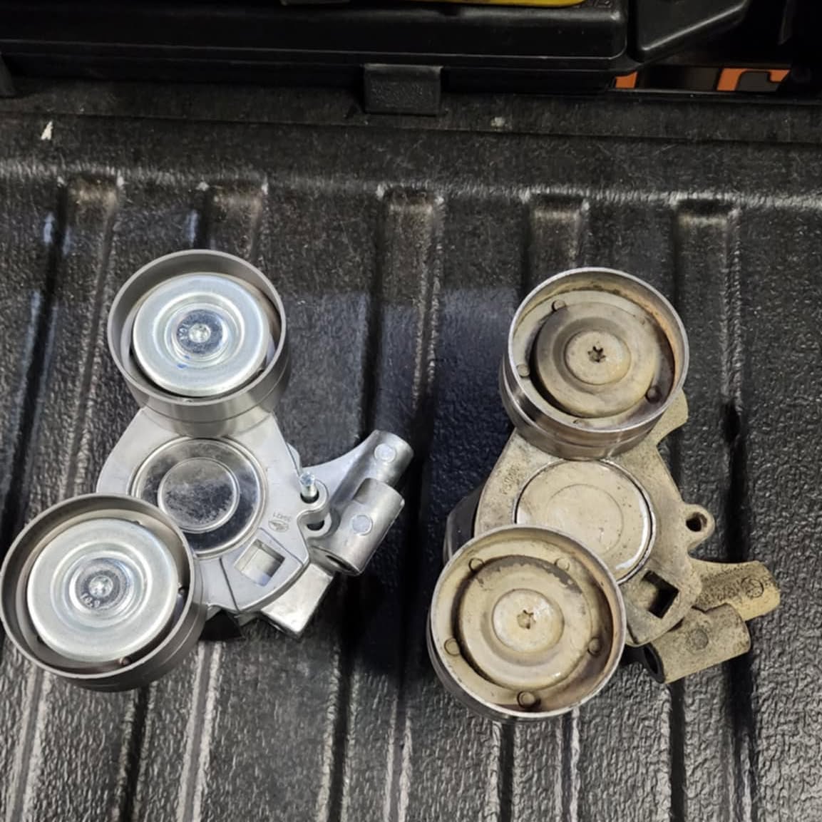 A new belt tensioner and pulley assembly beside the worn original, laid out on a ute tray